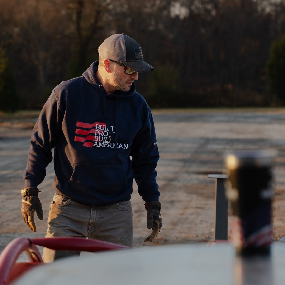 Man wearing a hoodie with text, standing next to a car in an open area with trees in the background.